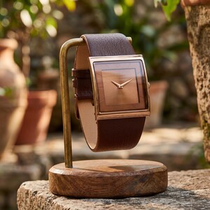 May include: A brown leather watch with a square face and gold-coloured trim is displayed on a wooden stand. The watch has a brown leather strap. The background features a blurred outdoor setting with potted plants.