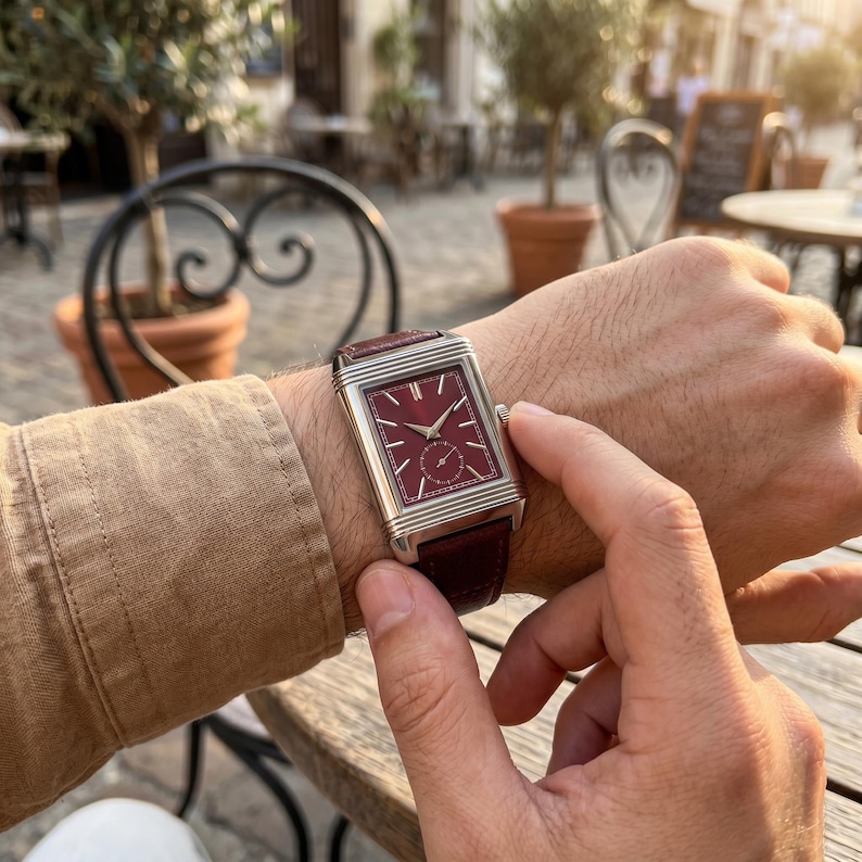 May include: A rectangular wristwatch with a burgundy face and a brown leather band. The watch has a silver-colored case and a small seconds subdial. The watch is being worn on a wrist.