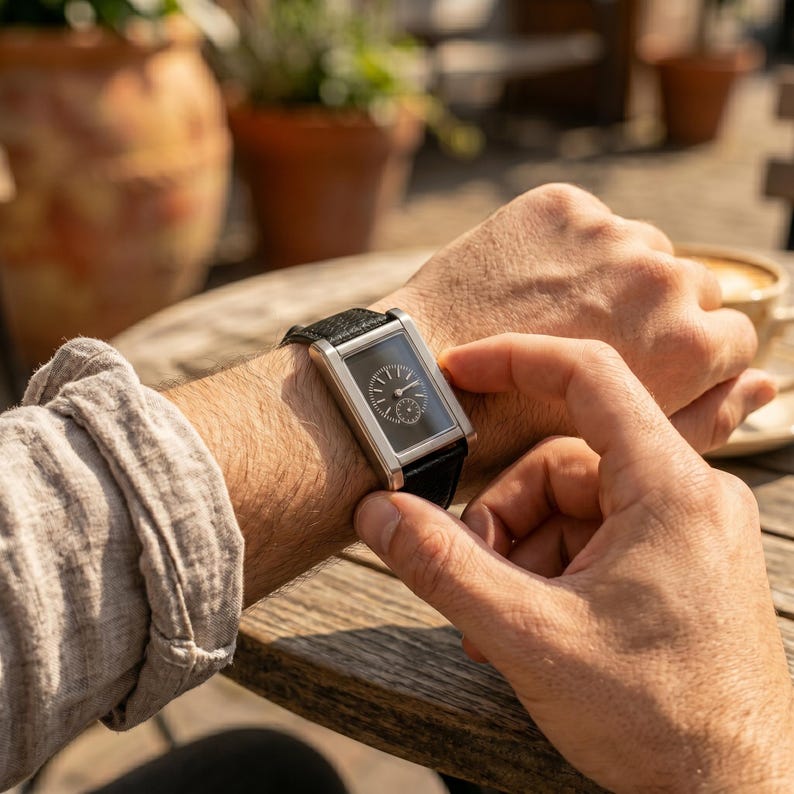May include: A rectangular wristwatch with a black face and silver-colored frame. The watch has a black leather band. The watch is on a person's wrist, with their hand adjusting the watch. The background includes a wooden table and a cup of coffee.