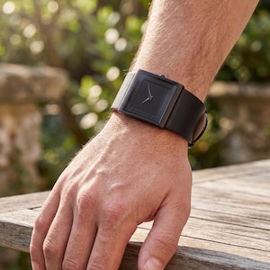 May include: A black wristwatch with a square face and a black leather strap. The watch has a minimalist design with silver hands. The watch is shown on a person's wrist. The strap is approximately 20 cm in length.