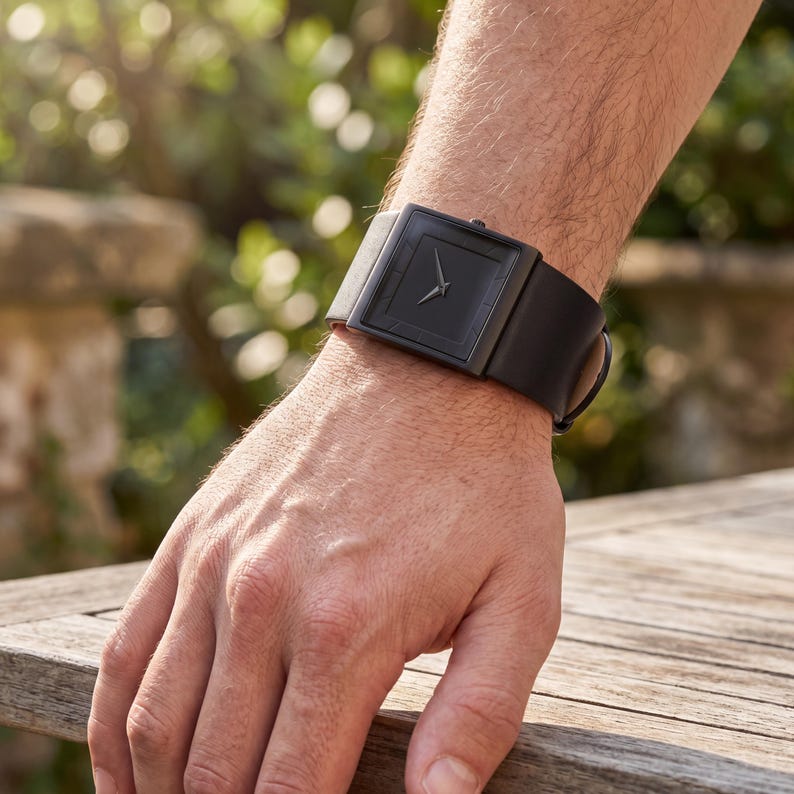 May include: A black wristwatch with a square face and a black leather strap. The watch has a minimalist design with silver hands. The watch is shown on a person's wrist. The strap is approximately 20 cm in length.