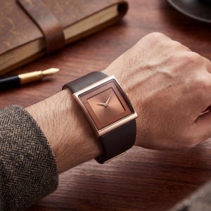 May include: A square-faced watch with a copper-coloured dial and rose gold-coloured frame. The watch has a dark brown leather strap. The watch is on a person's wrist.