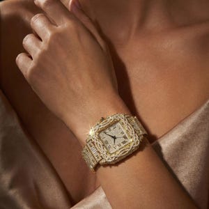 Op de afbeelding: Een gouden polshorloge met een vierkante wijzerplaat en een met diamanten bezette lunette en band. De wijzerplaat van het horloge heeft Romeinse cijfers en zwarte wijzers. Het horloge wordt om de pols gedragen.