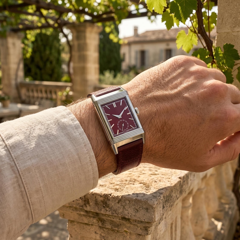 May include: A rectangular wristwatch with a burgundy face and silver-colored casing. The watch has a burgundy leather band. The watch is on a wrist, with a light-colored long-sleeve shirt visible.