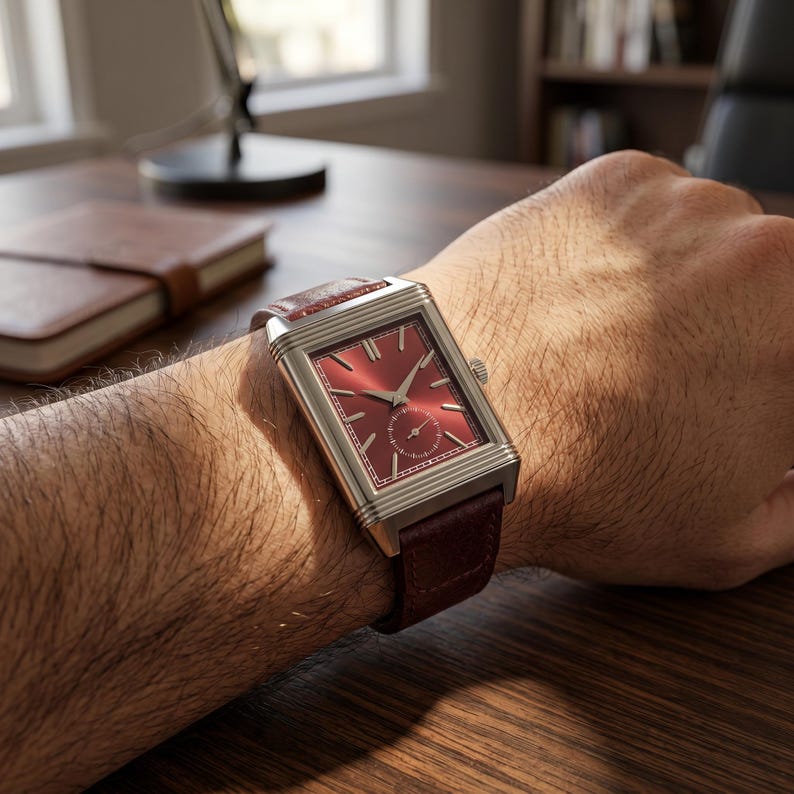 May include: A rectangular wristwatch with a red dial and silver-colored case is displayed on a wrist. The watch has a small seconds subdial and a dark red leather strap. The watch is a luxury item.