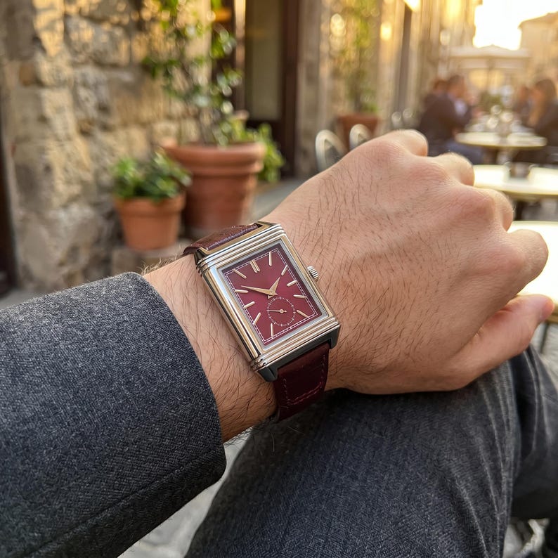 May include: A rectangular wristwatch with a burgundy face and a brown leather band. The watch has a silver-colored case and a small subdial. The watch is on a wrist, with a gray jacket sleeve visible.
