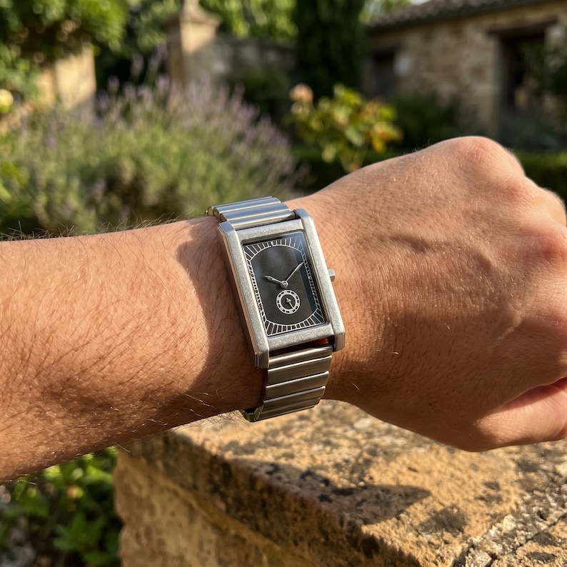 May include: A rectangular silver wristwatch with a black face. The watch has a silver band and a minimalist design. The watch is on a wrist, with a blurred outdoor background.