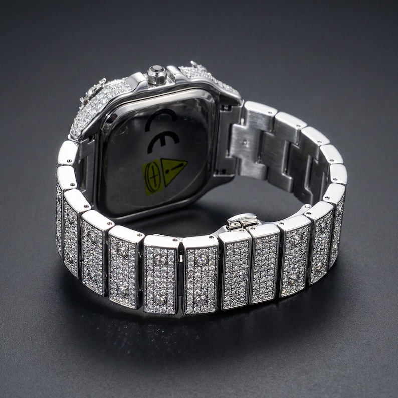 Op de afbeelding: Een zilverkleurig polshorloge met een vierkante wijzerplaat en een bijpassende band. Het horloge en de band zijn bezet met kleine, glinsterende steentjes. Het horloge wordt tegen een donkere achtergrond getoond.