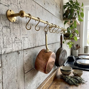 Könnte beinhalten: Eine horizontale Küchenstange aus Messing, die an der Wand montiert ist und mehrere S-Haken aus Messing aufweist. Ein runder Kupfertopf und eine dunkle Metallpfanne hängen an den Haken. Die Stange hat an beiden Enden runde Befestigungsplatten.
