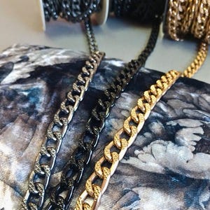 May include: Close-up of three metal chain rolls in silver, black, and gold. The chains are thick, with a curb link design. The chains are displayed on a patterned fabric background, with the rolls of chain in the background.
