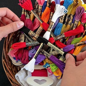 Op de afbeelding: Een verzameling meerkleurige strengen borduurgaren in een gevlochten bruine mand. Elke streng is voorzien van een zwart papieren label met de gouden tekst DMC 25. Het assortiment bevat levendige tinten rood, blauw, geel, paars, oranje en wit.