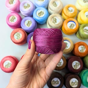 Könnte beinhalten: Eine Hand hält eine lebendige magentafarbene Garnspule, die ihre eng gewickelte Textur zeigt. Der Hintergrund zeigt eine Sammlung farbenfroher Garnspulen in Rosa-, Blau-, Gelb- und Brauntönen, ideal für Bastel- und Stickprojekte.