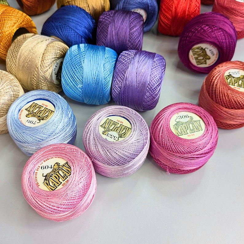 May include: A collection of small, round balls of polyester thread in various vibrant colors including pink, purple, blue, gold, and red. Each ball features a central white label with the brand name KAPLAN and a tiger illustration printed on it.