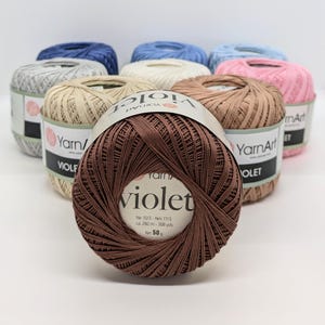 Könnte beinhalten: Eine Auswahl an YarnArt Violet Garnknäueln in verschiedenen Farben, darunter Braun, Rosa, Blau und Beige. Das braune Knäuel ist im Vordergrund, mit dem Wort "Violet" auf dem Etikett. Jedes Knäuel enthält ca. 282 Meter Garn, Nettogewicht 50 g.