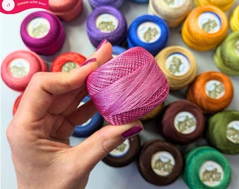 May include: A collection of small, round balls of embroidery thread in a wide variety of vibrant colors, including pink, blue, yellow, green, and brown. Each ball features a central white label with black text and a small graphic illustration.
