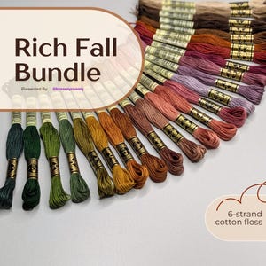 May include: A collection of 6-strand cotton embroidery floss skeins arranged in a fan shape, featuring a gradient of autumn-inspired colours including deep greens, golden yellows, burnt oranges, warm browns, and muted purples with gold labels.