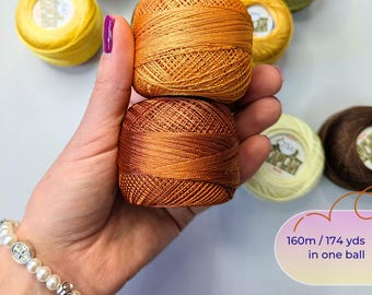 May include: Two balls of embroidery thread in shades of golden yellow and copper brown are held together. Each ball contains 174 yards of thread. Additional balls of thread in yellow, green, and brown are visible in the background.