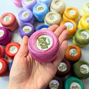 May include: A pink ball of Kaplan No. 50 crochet thread held in the palm, surrounded by numerous other colorful balls of thread. Each ball features a central white label with the brand name Kaplan, a tiger illustration, and a color number.