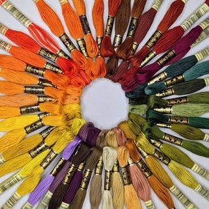 May include: A circular arrangement of DMC embroidery floss skeins in a spectrum of colors including orange, red, purple, green, yellow, and brown. Each skein features a paper label with a barcode and color number, arranged in a color wheel pattern.