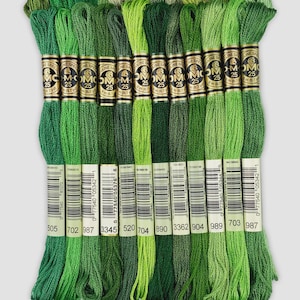 Op de afbeelding: Een verzameling groene borduurgaren in diverse tinten, van licht tot donker. Elk streng is genummerd en bevat merkinformatie. De garens zijn netjes gerangschikt, met een scala aan groene tinten.
