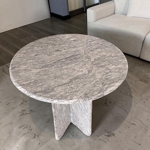 Può includere: Un tavolino rotondo con piano in marmo grigio e un'esclusiva base a tre gambe. Il tavolo è appoggiato su un tappeto grigio, con un divano bianco sullo sfondo. Il diametro del tavolo è di circa 100 cm.
