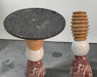 Puede incluir: Un pedestal de piedra con una superficie circular gris oscuro y una base compuesta por segmentos de mármol apilados en amarillo estriado, blanco esférico y granate cónico, junto a un candelabro alto a juego con componentes de mármol similares.