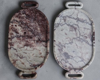 Puede incluir: Dos bandejas ovaladas de mármol con asas integradas en la parte superior e inferior. Una bandeja presenta vetas de color burdeos intenso y crema, mientras que la otra muestra patrones de mármol gris claro y blanco con un acabado superficial pulido.