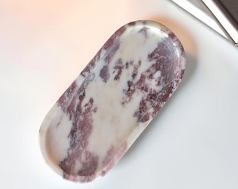 Puede incluir: Una bandeja de forma ovalada hecha de piedra pulida con un patrón de mármol natural con vetas de color burdeos intenso y blanco. La bandeja tiene una superficie lisa ligeramente hundida y bordes redondeados, mostrando las variaciones de color.