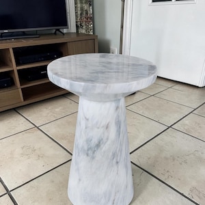 Puede incluir: Una mesa auxiliar cil&iacute;ndrica con una superficie lisa y pulida con un patr&oacute;n de m&aacute;rmol blanco y gris. La mesa tiene una parte superior circular y una base c&oacute;nica que se ensancha hacia el suelo, creando una estructura s&oacute;lida de piedra.