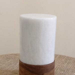 Pode incluir: Lâmpada cilíndrica de mármore branco com base de madeira castanha escura. A lâmpada tem um design simples e elegante, perfeito para uma mesa de cabeceira ou secretária. A lâmpada está sobre uma superfície de serapilheira.