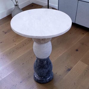 May include: A small, round side table with a white marble top. The base is made up of beige, white, and dark blue sections. The table is on a wooden floor. The table's height is approximately 60 cm.