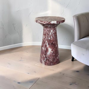 Puede incluir: Una mesa auxiliar cil&iacute;ndrica hecha de m&aacute;rmol pulido rojo y blanco. La mesa presenta una superficie superior circular que se transforma en una base c&oacute;nica tipo pedestal, mostrando las vetas naturales y variaciones de color de la piedra.