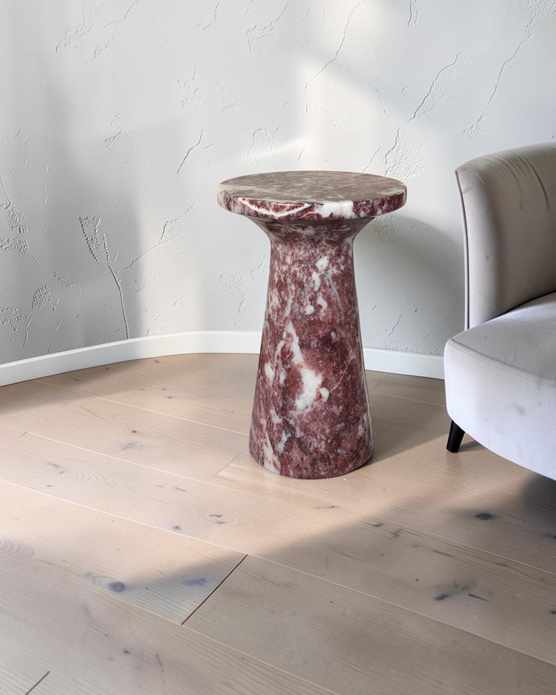 Puede incluir: Una mesa auxiliar cil&iacute;ndrica hecha de m&aacute;rmol pulido rojo y blanco. La mesa presenta una superficie superior circular que se transforma en una base c&oacute;nica tipo pedestal, mostrando las vetas naturales y variaciones de color de la piedra.