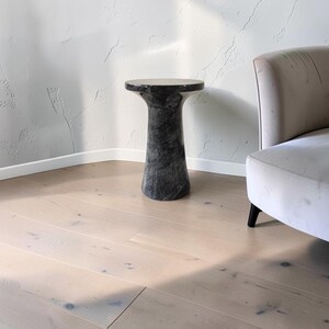 Puede incluir: Una mesa auxiliar cil&iacute;ndrica hecha de m&aacute;rmol gris oscuro con vetas blancas prominentes. La mesa presenta una superficie superior circular lisa que se estrecha hacia el centro antes de ensancharse ligeramente en la base, creando una forma de pedestal.