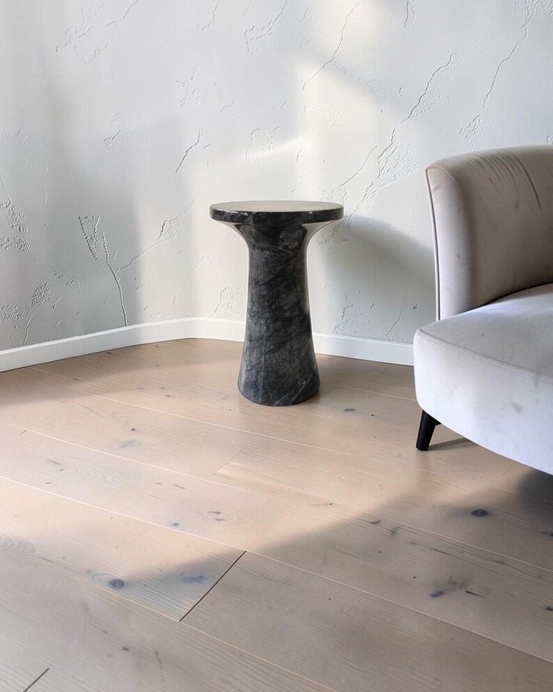 Puede incluir: Una mesa auxiliar cil&iacute;ndrica hecha de m&aacute;rmol gris oscuro con vetas blancas prominentes. La mesa presenta una superficie superior circular lisa que se estrecha hacia el centro antes de ensancharse ligeramente en la base, creando una forma de pedestal.