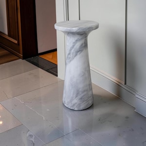 Puede incluir: Una mesa pedestal cil&iacute;ndrica fabricada en m&aacute;rmol blanco con vetas grises prominentes. La pieza presenta una superficie superior circular lisa que se transforma en una base c&oacute;nica y ensanchada, creando una estructura s&oacute;lida y estable.
