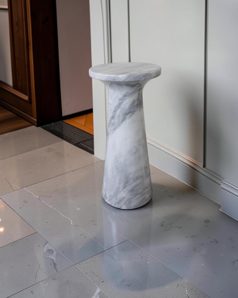 Puede incluir: Una mesa pedestal cil&iacute;ndrica fabricada en m&aacute;rmol blanco con vetas grises prominentes. La pieza presenta una superficie superior circular lisa que se transforma en una base c&oacute;nica y ensanchada, creando una estructura s&oacute;lida y estable.