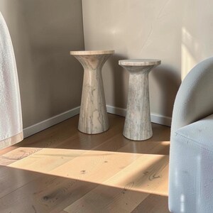 Puede incluir: Dos mesas auxiliares cil&iacute;ndricas hechas de piedra beige claro con vetas grises naturales. Cada mesa presenta una parte superior ensanchada y una base c&oacute;nica, con diferentes alturas. Las superficies de piedra tienen un acabado liso y pulido.
