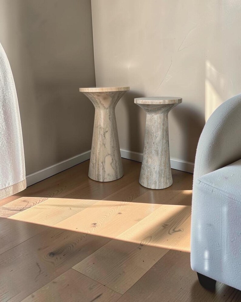 Puede incluir: Dos mesas auxiliares cil&iacute;ndricas hechas de piedra beige claro con vetas grises naturales. Cada mesa presenta una parte superior ensanchada y una base c&oacute;nica, con diferentes alturas. Las superficies de piedra tienen un acabado liso y pulido.