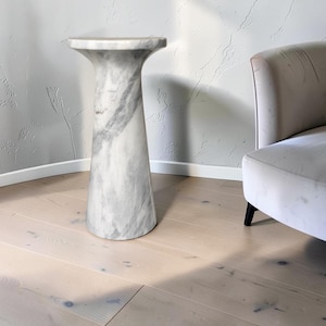 Puede incluir: Una mesa pedestal de m&aacute;rmol blanco con una base cil&iacute;ndrica lisa que se ensancha en la parte superior para formar una superficie circular. La piedra presenta vetas grises naturales, creando un patr&oacute;n veteado en toda la estructura.