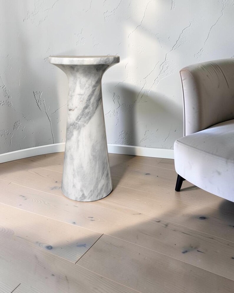 Puede incluir: Una mesa pedestal de m&aacute;rmol blanco con una base cil&iacute;ndrica lisa que se ensancha en la parte superior para formar una superficie circular. La piedra presenta vetas grises naturales, creando un patr&oacute;n veteado en toda la estructura.