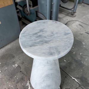 Puede incluir: Una mesa auxiliar redonda hecha de m&aacute;rmol blanco con vetas grises. La mesa presenta una superficie superior circular lisa y una base de pedestal s&oacute;lida y c&oacute;nica que se ensancha hacia el suelo, mostrando una textura de piedra pulida uniforme.