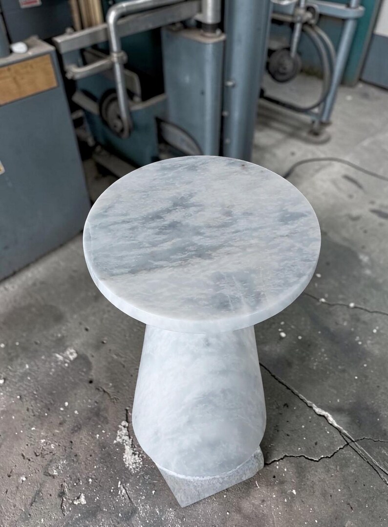 Puede incluir: Una mesa auxiliar redonda hecha de m&aacute;rmol blanco con vetas grises. La mesa presenta una superficie superior circular lisa y una base de pedestal s&oacute;lida y c&oacute;nica que se ensancha hacia el suelo, mostrando una textura de piedra pulida uniforme.