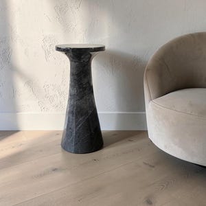 Puede incluir: Una mesa auxiliar alta y cil&iacute;ndrica hecha de m&aacute;rmol gris oscuro con vetas blancas prominentes. La mesa presenta una superficie lisa y pulida con una parte superior ensanchada, apoyada sobre una base ancha y estable que se estrecha hacia el centro.