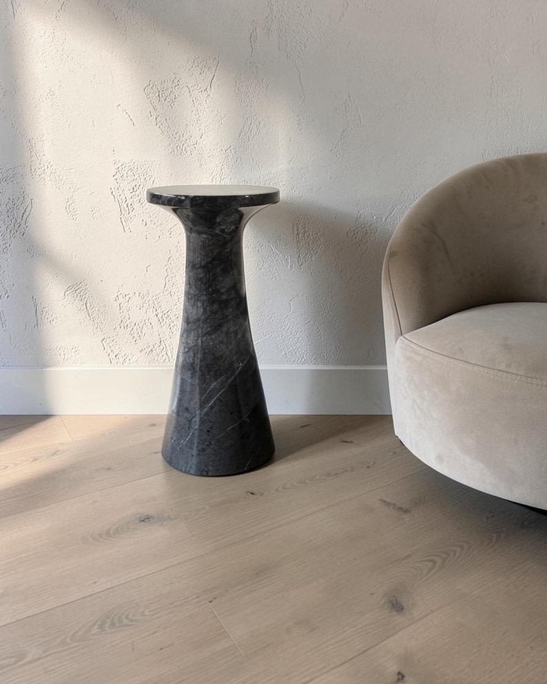 Puede incluir: Una mesa auxiliar alta y cil&iacute;ndrica hecha de m&aacute;rmol gris oscuro con vetas blancas prominentes. La mesa presenta una superficie lisa y pulida con una parte superior ensanchada, apoyada sobre una base ancha y estable que se estrecha hacia el centro.