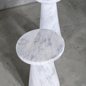 Puede incluir: Dos mesas auxiliares de m&aacute;rmol blanco con tablero circular y base c&oacute;nica estrecha. La piedra presenta vetas grises naturales en toda la superficie, creando un acabado liso y pulido tanto en el tablero como en la estructura del pedestal.