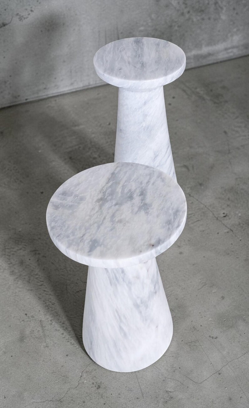Puede incluir: Dos mesas auxiliares de m&aacute;rmol blanco con tablero circular y base c&oacute;nica estrecha. La piedra presenta vetas grises naturales en toda la superficie, creando un acabado liso y pulido tanto en el tablero como en la estructura del pedestal.