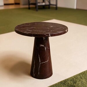 Puede incluir: Una mesa auxiliar redonda hecha de m&aacute;rmol marr&oacute;n oscuro con vetas blancas prominentes. La mesa presenta una superficie circular plana sostenida por una base c&oacute;nica &uacute;nica, s&oacute;lida y estrecha que se ensancha hacia el suelo, creando un pedestal estable.