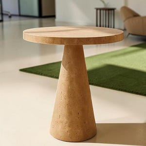 Puede incluir: Una mesa auxiliar redonda hecha de piedra porosa de color tostado claro. La mesa presenta una superficie superior circular plana apoyada por una base c&oacute;nica &uacute;nica y s&oacute;lida que se estrecha hacia el suelo, mostrando un acabado natural y texturizado.