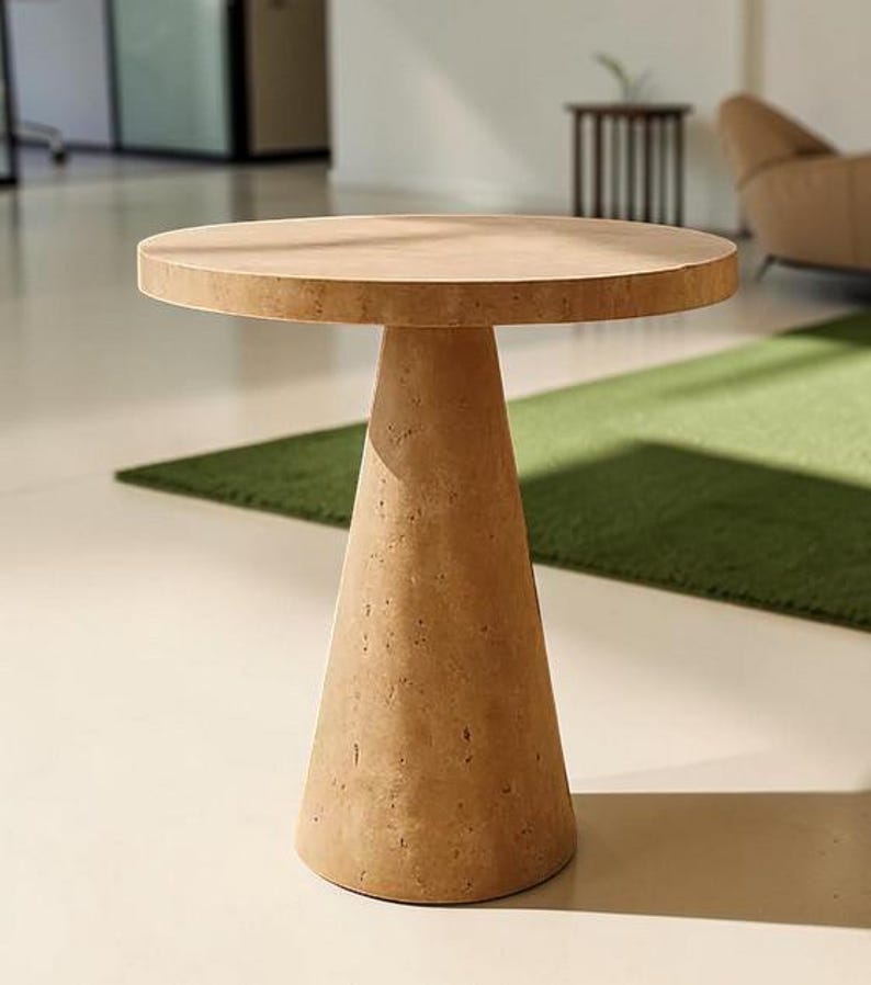 Puede incluir: Una mesa auxiliar redonda hecha de piedra porosa de color tostado claro. La mesa presenta una superficie superior circular plana apoyada por una base c&oacute;nica &uacute;nica y s&oacute;lida que se estrecha hacia el suelo, mostrando un acabado natural y texturizado.